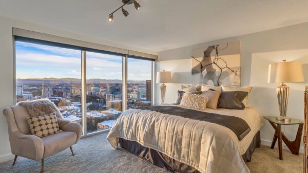 Bedroom with carpet, plush bed with headboard, lounge chair, and large windows overlooking Denver.