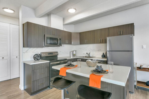 Kitchen with wood floor and cabinets, light counters, stainless steel appliances, and island with stools.
