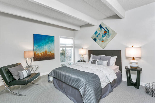 Bedroom with carpet, large bed with plush bedding, modern leather chair, artwork and large window.