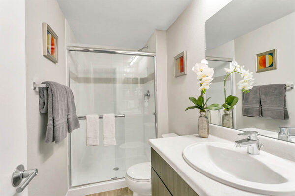 Bathroom with light counters, large mirror, tiled shower with glass door, and artwork.