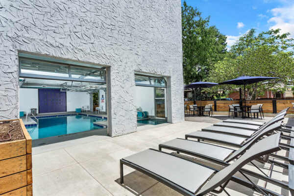 Outdoor patio with lounge chairs, tables with umbrellas, and garage doors to indoor pool.