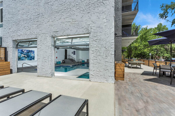 Outdoor patio with lounge chairs, tables with umbrellas, and garage doors to indoor pool.