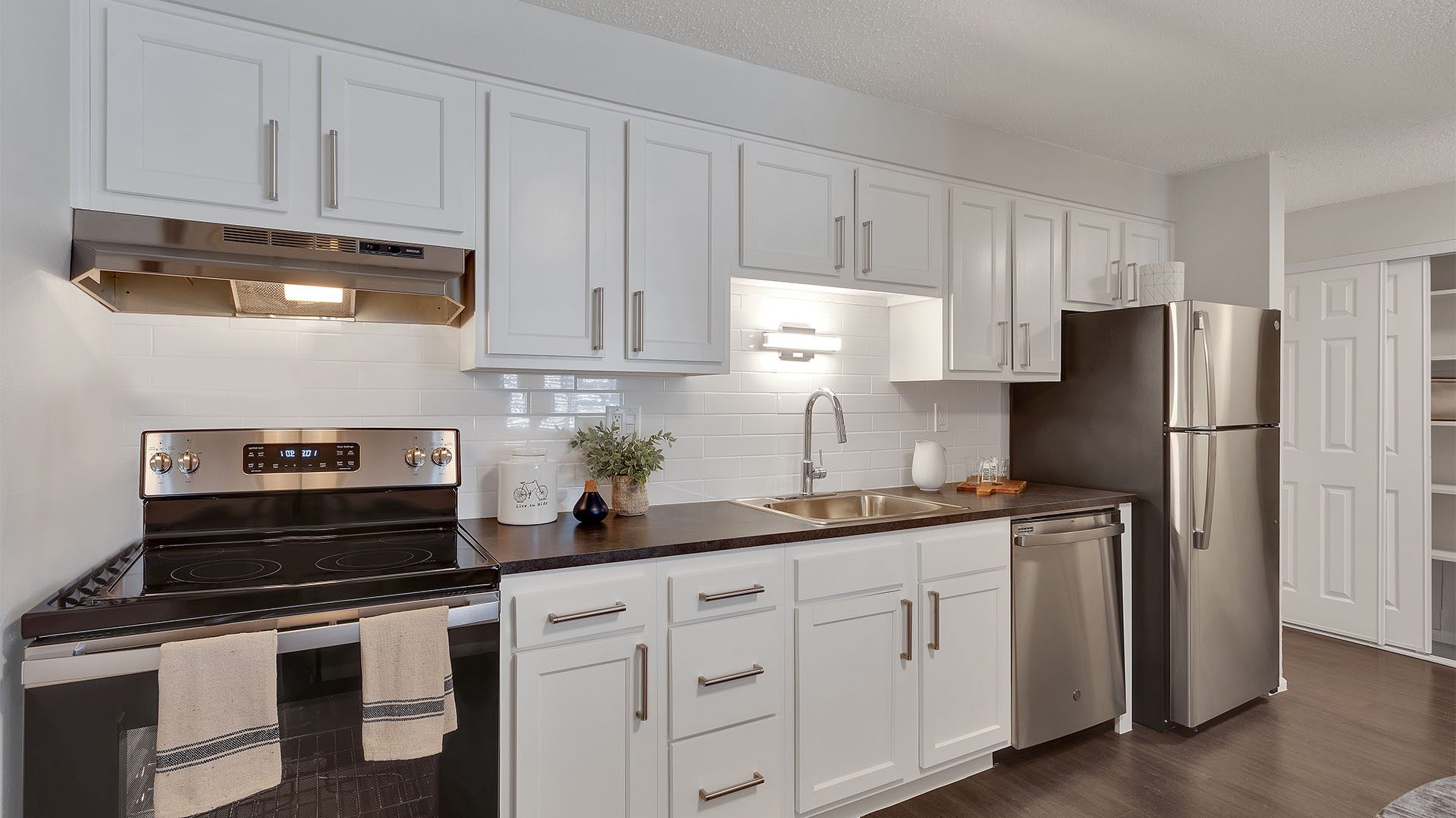 Kitchen with wood floor, white cabinets, dark counters, stainless steel appliances, and tiled backsplash.
