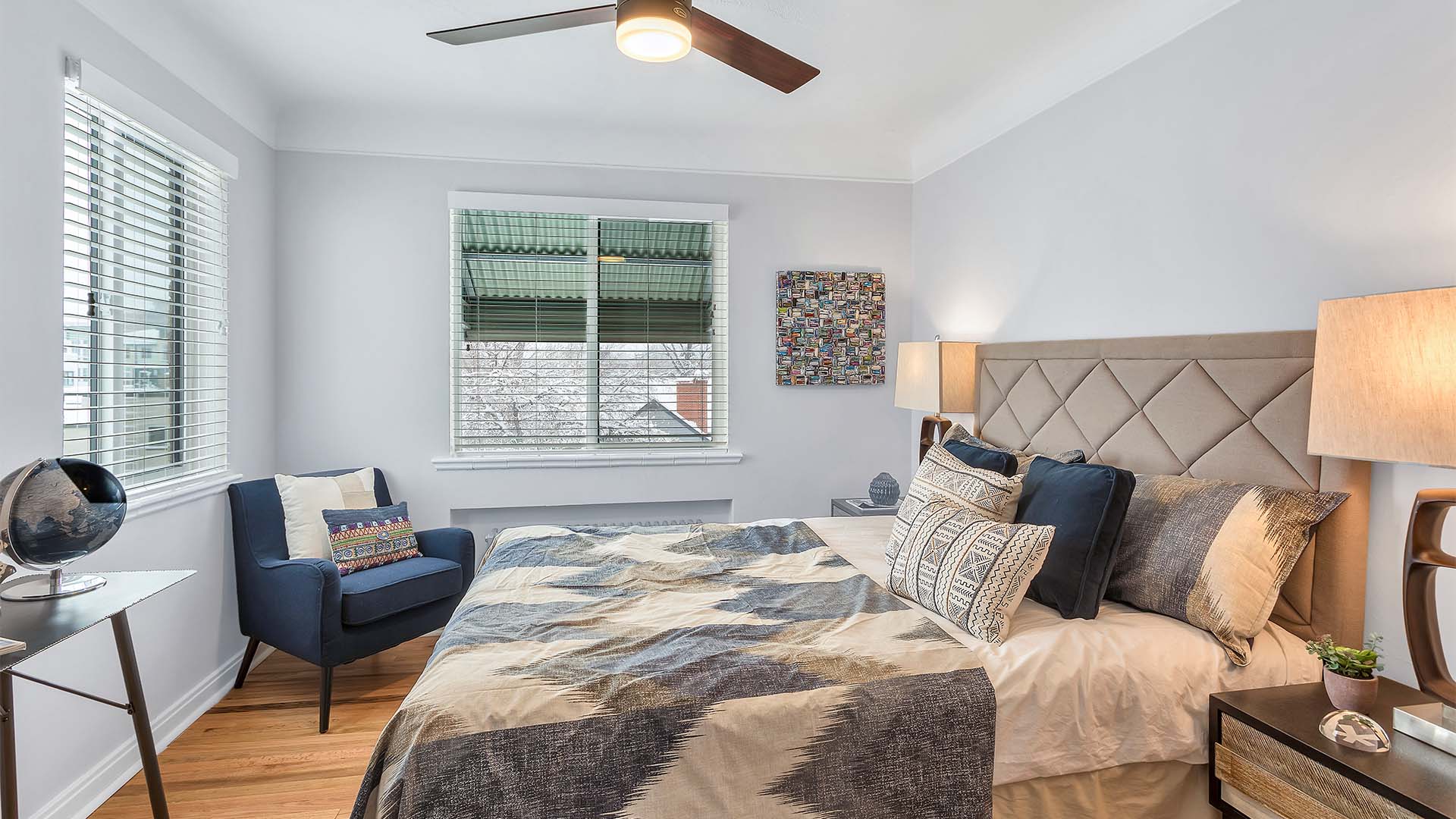 Bedroom with wood floor, plush bed with headboard, lounge chair, and large windows.