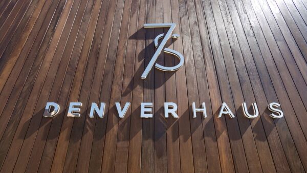 Detail of metallic 7 S Denver Haus monument sign on wood paneling in bright sunshine.