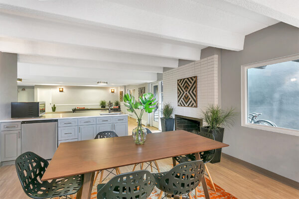 Community kitchen with island, wood table with modern chairs, large window, and brick accent wall with fireplace.