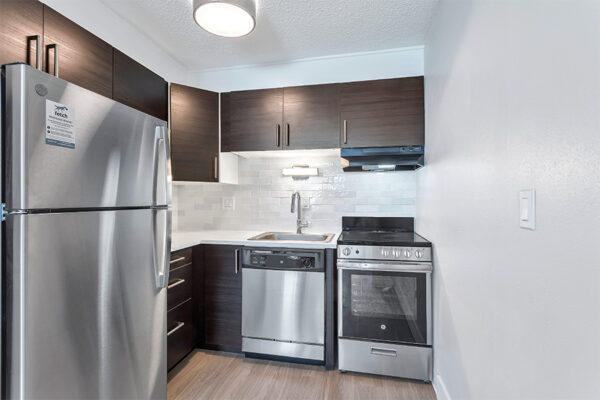 Kitchen with wood floor, dark cabinets, light counters, stainless steel appliances, and tiled backsplash.