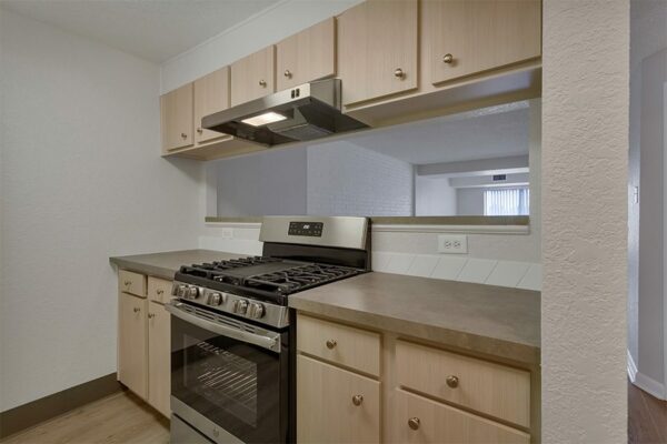 Kitchen with wood floor and cabinets, tan counters, stainless steel appliances, and opening to living room.