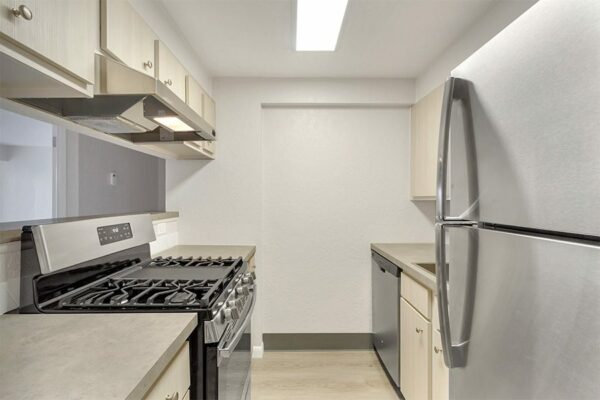 Kitchen with wood floor and cabinets, tan counters, stainless steel appliances, and opening to living area.