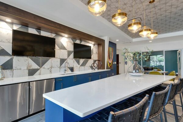 Community kitchen with blue cabinets, white counters, breakfast bar, wall mounted TVs, and decorative lighting fixtures.