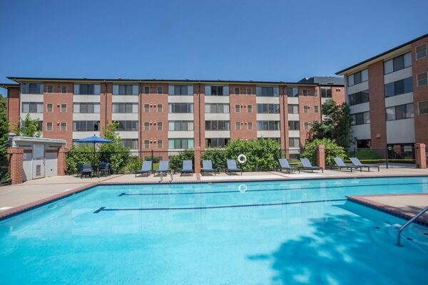 Fenced pool area with lounge chairs, tables with umbrellas, and tall trees.