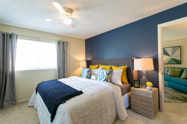Bedroom with carpet, blue accent wall, bed with plush headboard, large window, and door to living room.