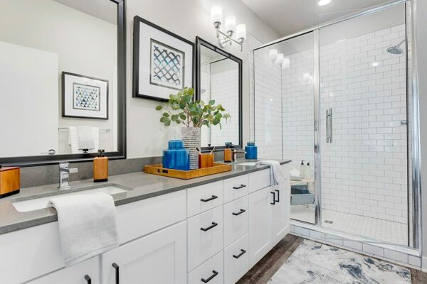 Bathroom with white cabinets, grey counters, two large framed mirrors, and tiled shower with glass door.