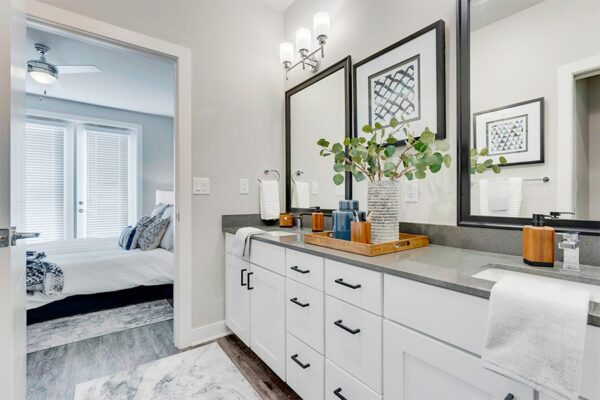 Bathroom with white cabinets, grey counters, framed mirrors, and door to bedroom.