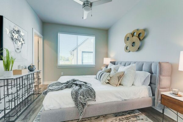 Bedroom with large bed with plush headboard, modern furniture and artwork, ceiling fan, and large window.