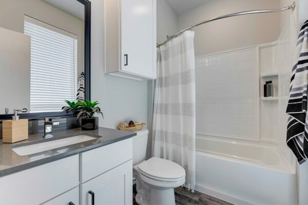 Bathroom with white cabinets, grey counters, large framed mirror, and tiled shower tub with curtain.