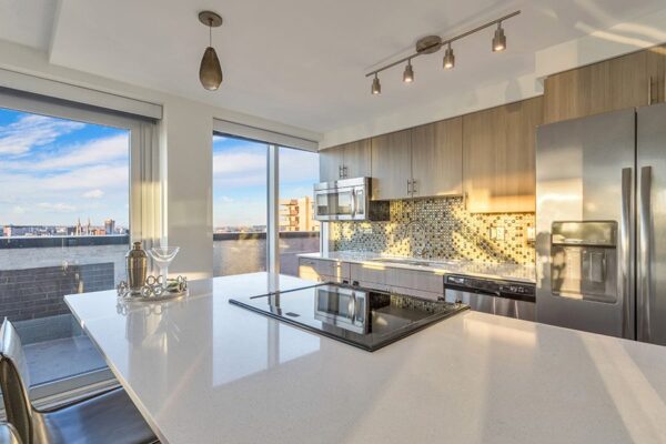 Apartment kitchen with wood floor and cabinets, white counters, stainless steel appliances, and tiled backsplash.