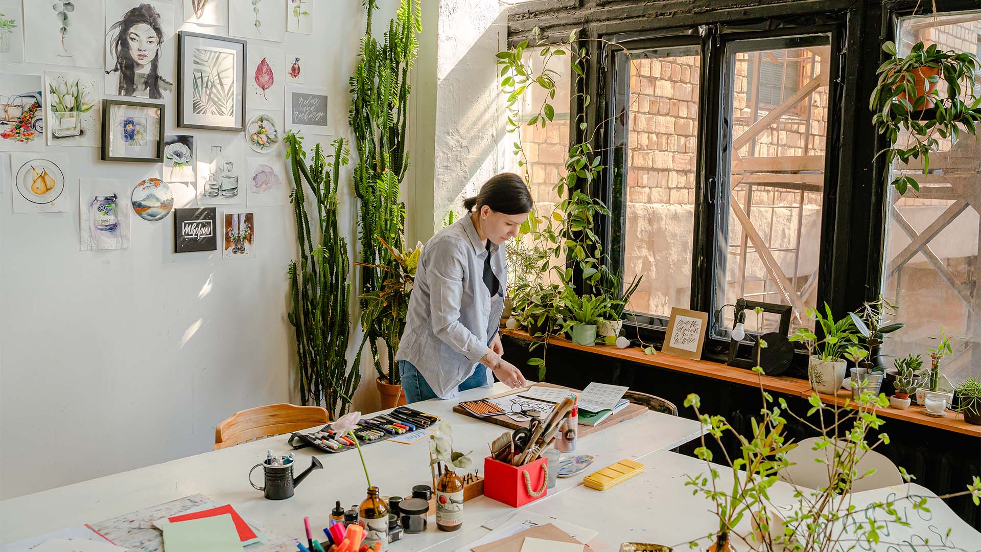 Artist in studio with plants, large windows, artwork, and art supplies.