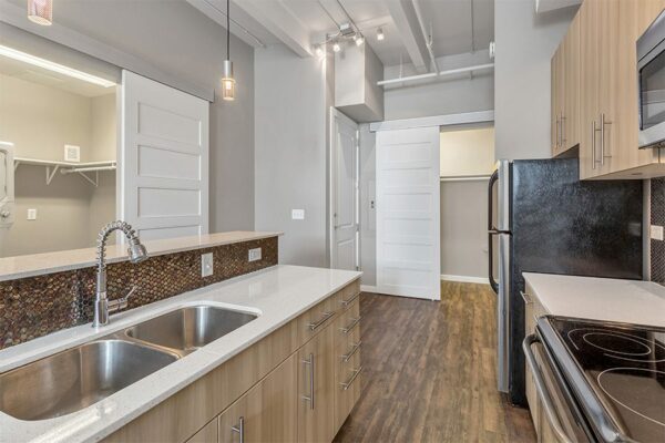Kitchen with wood floor and cabinets, light counters, stainless steel sink and faucet, and round tiled backsplash.