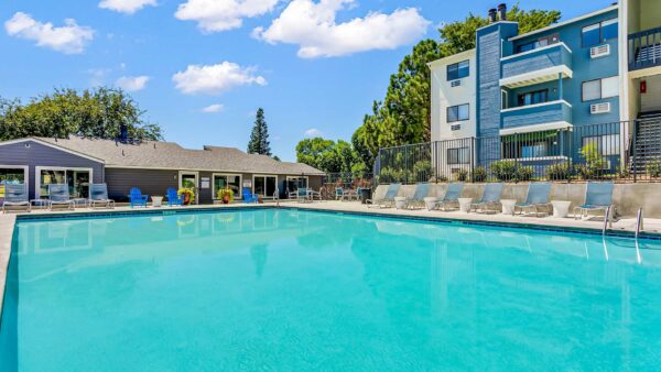 Fenced pool area with lounge chairs, dining tables, and trees overlooked by apartment balconies.