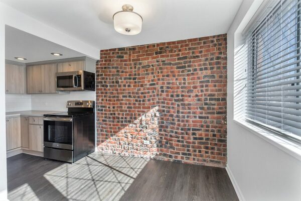 Living area and kitchen with wood floor, brick accent wall, wood cabinets, and stainless steel appliances.