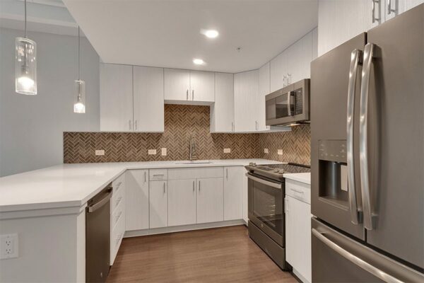 Kitchen with wood floor, light cabinets and counters, stainless steel appliances, tiled backsplash, and pendant lights.