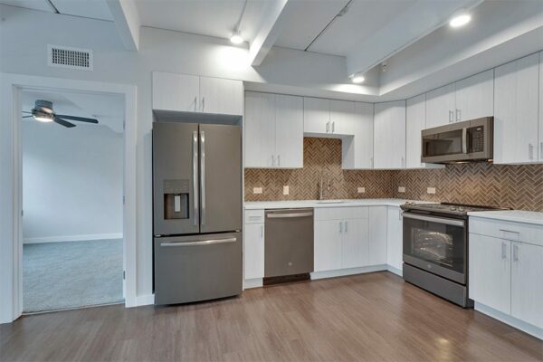 Kitchen with light cabinets and counters, stainless steel appliances, tiled backsplash, and door to bedroom.