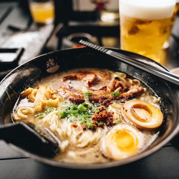 Bowl of ramen with meat, noodles, and eggs on table with beer.