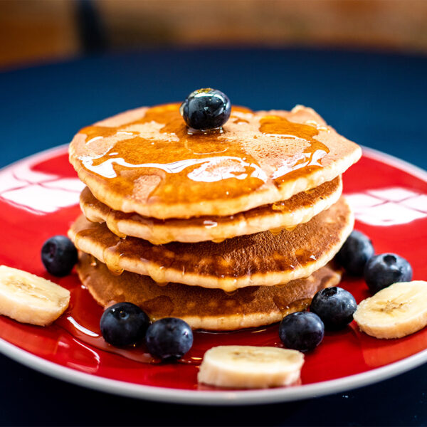Tall stack of pancakes with syrup, blueberries and bananas on red plate.