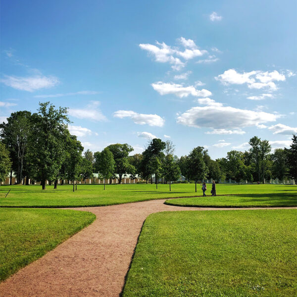 Park with dirt walkways, trimmed grass, and tall trees under cloudy blue skies.