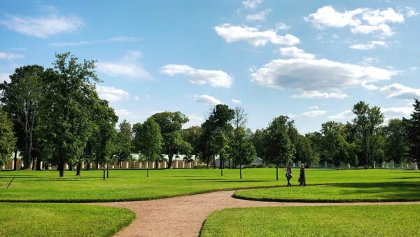 Park with dirt walkways, trimmed grass, and tall trees under cloudy blue skies.