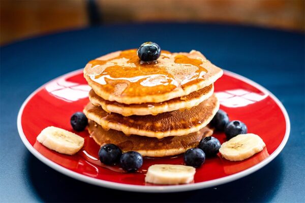 Tall stack of pancakes with syrup, blueberries and bananas on red plate.