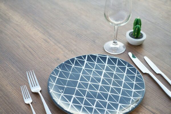 Place setting with geometric blue plate, silverware, wine glass, and cactus on wood table.
