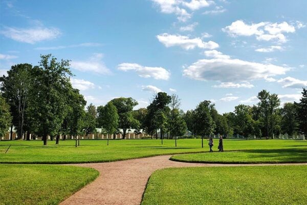 Park with dirt walkways, trimmed grass, and tall trees under cloudy blue skies.