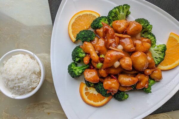 Orange chicken with broccoli on white plate with bowl of white rice.